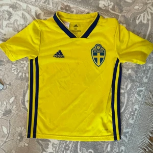 Svenska landslagets fotbollströja XS - Gul fotbollströja från Adidas med blå detaljer och Sveriges landslagsemblem på bröstet. Klassisk V-ringning med blå kant, tre blå ränder längs sidorna och 'Sverige' tryckt i nacken. Tillverkad i lätt och ventilerande Climalite-material.den har liten föd färg som ni kan se i bild nr 4 och liten riven som ni ser på sista bild .barn storlek är den som en 8-9 år 