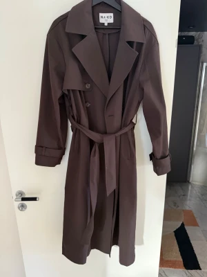 Brun trenchcoat från NA-KD - Snygg brun trenchcoat från NA-KD med klassisk dubbelknäppning, långa ärmar och bälte i midjan. Storlek 36