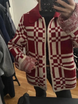Rödvit stickad kofta med rutmönster - Säljer en unik stickad kofta i rött och vitt med rutigt mönster. Koftan har stora fickor framtill, bred krage och knappar längs hela framsidan. Materialet ser ut att vara mjukt och tjockt, perfekt för lager på lager. Snygg retrovibe och passar till många outfits.💕