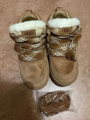 Ugg lowmel - Snygga beige sneakers från UGG med vit fuskpäls på insidan och grova snören i beige och vitt mönster. Skorna har en robust platt sula, rund tå och detaljer i mocka och textil. Perfekta för dig som vill ha både stil och komfort.