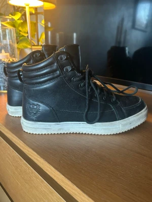 Svarta kängor i skinn med snörning - Säljer ett par svarta kängor i skinn med vit sula och snörning. Skorna har en vadderad kant runt ankeln och en diskret logga på sidan. Använt två sesonger 