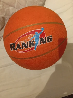 Basketboll Super Ranking orange - Säljer en klassisk orange basketboll från Super Ranking med guldfärgade linjer och tryckt logga med en blå siluett av en basketspelare. Bollen är tillverkad i slitstarkt gummi och har tydlig textur för bra grepp. Perfekt för spel på både ute- och inomhusplan. (BOLLEN ÄR INTE PUMPAD)
