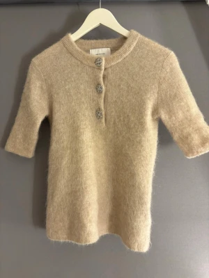 Beige stickad tröja H&M Edition - Beige stickad tröja från H&M Edition med halvlånga ärmar och tre dekorativa knappar framtill. Tröjan har en mjuk, fluffig känsla och rund halsringning. Perfekt för lager-på-lager och enkel att matcha med olika outfits.