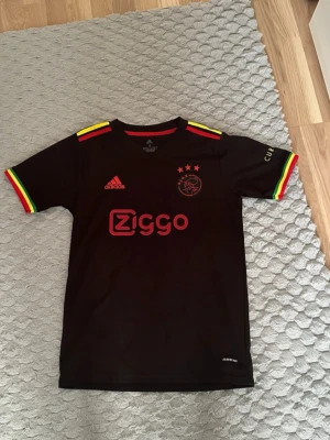 Ajax svart matchtröja Antony #11 - Säljer en svart Ajax matchtröja från Adidas med Ziggo-logga och röda detaljer framtill. Tröjan har gula, röda och gröna ränder på ärmarna samt namn och nummer 'Antony 11' i gult på ryggen. Korta ärmar och klubbmärke på bröstet. Perfekt för fotbollsfans!