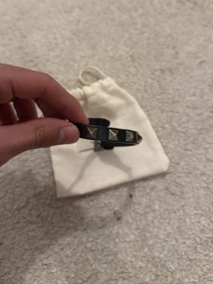 Valentino svart läderarmband nitar - Snyggt svart armband i läder från Valentino Garavani med silverfärgade pyramidnitar och spänne. Armbandet har en stilren och edgy design som passar perfekt till din outfit. Levereras med originalpåse.