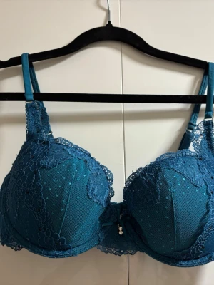 Blå spets-bh med rosett och pärla - Snygg blå bh med vadderade kupor, spetsdetaljer och små prickar i tyget. Mellan kuporna sitter en liten rosett och ett hängande pärlsmycke. Axelbanden är justerbara och bh:n har en feminin och elegant look.