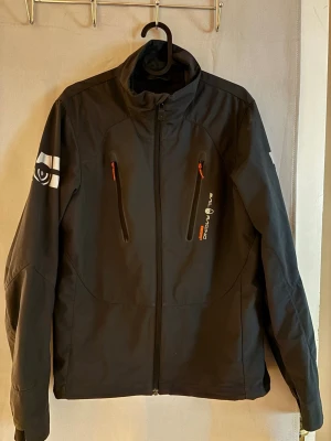 Svart vindjacka från Sail Racing - Svart vindjacka från Sail Racing med hög krage, dragkedja framtill och två bröstfickor med orange detaljer. Jackan har logotyp på bröstet och ärmen samt tryck på ryggen. Perfekt för blåsiga dagar och sportiga outfits.