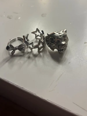 Tre silverfärgade ringar med motiv - Säljer tre unika ringar i silverfärg: en med rosett, en med stjärnor och en med ett detaljerat tigerhuvud. Perfekta för dig som vill sticka ut och addera lite edge till din stil. Alla ringar är i metall och har coola, ungdomliga designer.