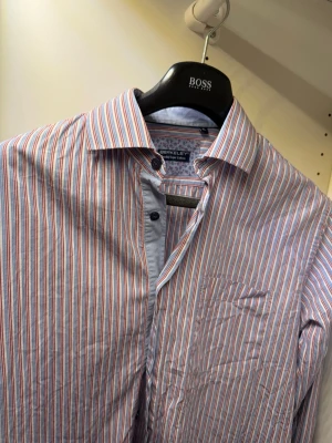 Randig skjorta från Berkeley i bomull - Säljer en snygg randig skjorta från Berkeley i Twofold Super Cotton. Skjortan har tunna ränder i rött, blått och vitt, klassisk krage och bröstficka. Perfekt för dig som gillar stilrena och färgglada skjortor.