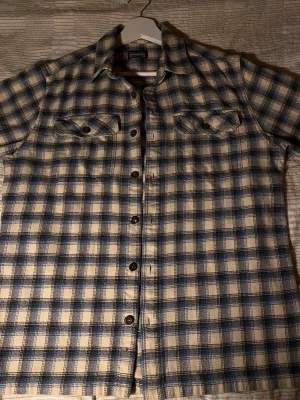 Rutig skjorta från Patagonia i bomull - Säljer en rutig skjorta från Patagonia i ekologisk bomull. Skjortan har blå och beige rutor, två bröstfickor med knappar och klassisk krage. Perfekt för dig som gillar avslappnad stil och vill ha något bekvämt och snyggt. Storlek S
