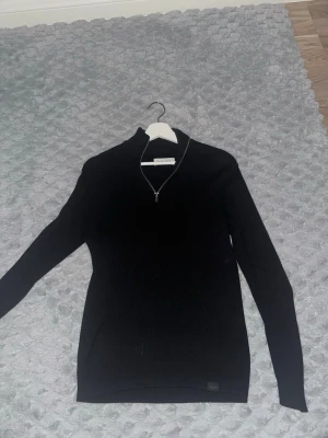 Svart stickad tröja från Pier One - Snygg svart stickad tröja från Pier One med ribbad struktur och half zip-dragkedja vid halsen. Tröjan har lång ärm och en klassisk, 