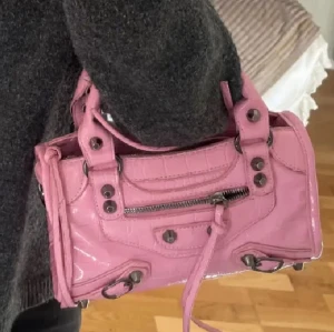 Rosa handväska med dragkedja - Cool rosa handväska med glansig finish och snygga metalldetaljer. Väskan har en dragkedja framtill, dekorativa nitar och dubbla handtag. Perfekt accessoar för att lyfta din outfit.