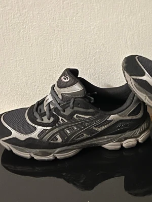 Svarta Asics sneakers med grå detaljer - Snygga svarta Asics sneakers med grå detaljer och meshpaneler för extra andningsförmåga. Skorna har klassisk snörning och en robust sula som ger bra stöd. Perfekta för dig som gillar sportig stil och komfort. 