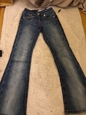 Blå bootcut jeans från Gina Tricot - Säljer ett par blå bootcut jeans från Gina Tricot med snygga slitningar och kontrastsömmar. Klassisk femficksmodell och knappgylf. Perfekta för dig som gillar en avslappnad men trendig look.