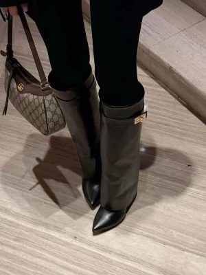 Svarta höga stövlar med spetsig tå - Säljer ett par svarta höga stövlar i skinn med spetsig tå och dekorativt guldfärgat spänne upptill. Stövlarna har en elegant och stilren design som passar perfekt till många outfits.