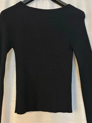 Svart ribbad topp med öppen rygg - Trendig svart ribbad topp med lång ärm och fyrkantig urringning. Toppen har en snygg öppen rygg med knytband upptill som ger en extra detalj. Perfekt för dig som gillar stilrena plagg med en twist.