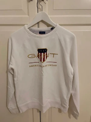 Vit sweatshirt från Gant - Vit sweatshirt från Gant med tryckt logga och sköld på bröstet i guld, marinblått och rött. Klassisk rund halsringning och ribbade muddar vid ärmslut och nederkant. Perfekt för en sportig och avslappnad stil.