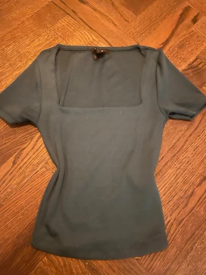 Mörkgrön topp med fyrkantig ringning - Stilren mörkgrön topp med fyrkantig ringning och korta ärmar. Toppen har en figurnära passform och är tillverkad i ett mjukt, stretchigt material som känns bekvämt mot huden. Perfekt att matcha med både jeans och kjol.