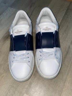 Valentino Garavani vita och blå sneakers äkta - Snygga vita sneakers från Valentino Garavani med breda mörkblå detaljer över mitten och klassiska nitar på hälen. Skorna har snörning och är tillverkade i skinn för en exklusiv känsla. Perfekta för dig som vill ha en stilren och modern look.alt till går,  Såklart äkta, priset går o diskutera 