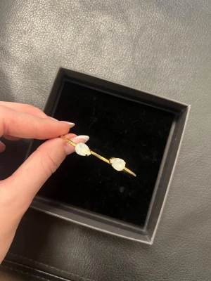 Guldigt armband med vita stenar - Snyggt och stilrent armband i guldfärgad metall med två stora, droppformade vita stenar i varje ände. Armbandet är öppet och justerbart, perfekt för att ge en elegant touch till din outfit. Kommer i en svart presentask.