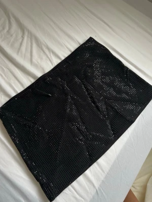 Svart glittrig kortkjol - Säljer en svart glittrig kortkjol med små paljetter över hela tyget. Kjolen har en rak passform och är perfekt för dig som vill sticka ut lite extra. Enkel att matcha med både toppar och boots.