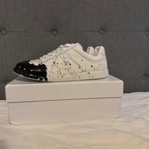 Maison margiela gats - Unika vita sneakers med svart färgstänksmönster över tå och sidor. Klassisk låg modell med snörning och rund tå. Tillverkade i syntetmaterial för en modern look. Perfekta för dig som vill sticka ut med din stil.