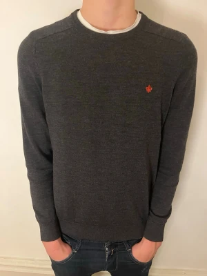 Mörkgrå tröja från Morris - Säljer en stilren mörkgrå tröja från Morris med rund halsringning och diskret röd logga broderad på bröstet. Tröjan har långa ärmar och är tillverkad i ett mjukt, tunt material som passar perfekt till lager på lager.