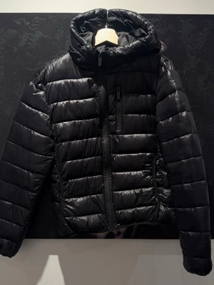 Svart pufferjacka från Closure London - Snygg svart pufferjacka från Closure London med huva och dragkedja framtill. Jackan har quiltad design, två sidofickor med dragkedja och en bröstficka. Perfekt för kalla dagar och ger en modern, sportig look.