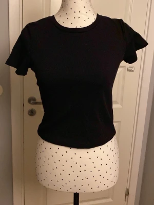 Svart ribbad t-shirt med volangärm - En svart ribbad t-shirt med rund hals och korta ärmar. T-shirten har en croppad passform och är tillverkad i mjuk bomull som känns skön mot huden. Perfekt för dig som gillar en enkel men ändå trendig stil.