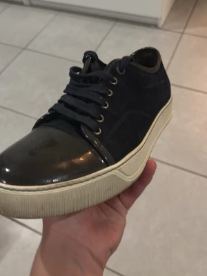 Lanvins - Stilrena svarta sneakers med snörning och vit sula. Ovandelen är i mocka och tån är lackad för en snygg kontrast. Skon har rund tå och klassisk låg profil som passar till många olika outfits. Ingen låda tillkommer.