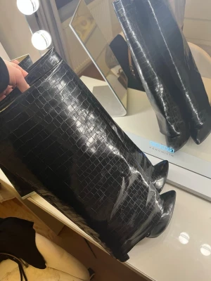 Svarta krokopräglade stövlar med klack - Säljer ett par svarta stövlar med krokodilpräglat mönster och hög klack. Stövlarna har spetsig tå och är tillverkade i glansigt syntetmaterial. Perfekta för att lyfta vilken outfit som helst.
