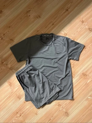 Grå Nike dry fit set - Snyggt grått träningsset från Nike med t-shirt och shorts. Båda plaggen är tillverkade i ett lätt och ventilerande material, perfekt för träning eller sport. Diskret Nike-logga på tröjan. Setet har en sportig och stilren look.