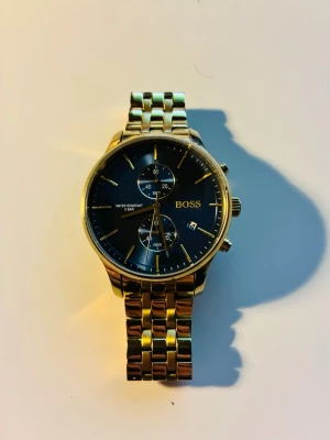 Guldfärgad klocka från BOSS - Stilren guldfärgad klocka från BOSS med blå  urtavla och guldiga detaljer. Klockan har länkarmband i metall, två subdials och datumvisning. Perfekt accessoar för dig som gillar klassisk och lyxig stil.  Klockan är som ny, använd max 5 gånger.