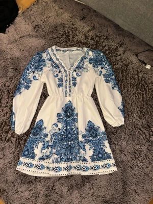 Vit och blå boho-klänning - Vit kortklänning med blått boho-inspirerat mönster. Klänningen har långärmade puffärmar, v-ringning med broderade detaljer och markerad midja. Perfekt för dig som gillar unika och färgstarka plagg.