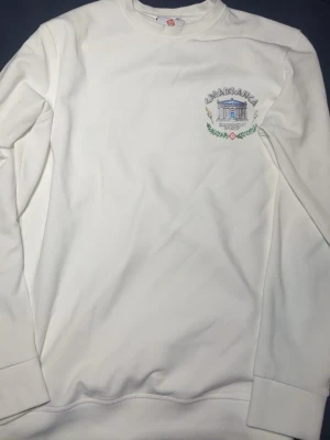 Vit sweatshirt från Casablanca - Säljer en vit sweatshirt från Casablanca med broderad logga och motiv på bröstet. Tröjan har rund hals, långa ärmar och ribbade muddar. Perfekt för dig som gillar stilrena och sportiga plagg med snygga detaljer.