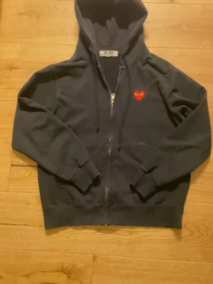 Svart hoodie från Comme des Garçons Play - Svart hoodie med dragkedja från Comme des Garçons Play. Klassisk design med huva och fickor framtill, samt det ikoniska röda hjärtat med ögon broderat på bröstet. Tillverkad i mjuk bomull för en skön känsla.