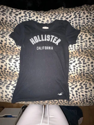 Svart Hollister t-shirt - Snygg svart t-shirt från Hollister med vit text 'Hollister California' på bröstet och liten logga nere vid fållen. Klassisk rund halsringning och korta ärmar. 