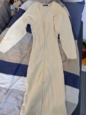 Lång cremevit klänning med knappar - Snygg långklänning i cremevitt med v-ringning och dekorativa knappar längs hela framsidan. Klänningen har lång ärm och är tillverkad i ett mjukt stickat material som känns skönt mot huden. Perfekt för dig som gillar stilrena och eleganta plagg. Lite slitet framme se på bilden, lagt till knapp går att ha kvar eller ta bort. 