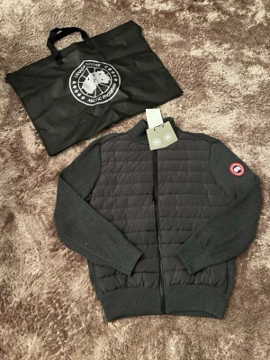 Canada Goose Cardigan - Snygg svart cardigan jacka från Canada Goose med quiltad front och stickade ärmar. Jackan har dragkedja framtill och klassisk logotyp på ärmen. Perfekt för lager-på-lager och stilrena outfits.