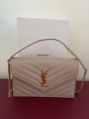 Beige quiltad kuvertväska YSL skinn - Supersnygg väska från YSL!! Baksidan har en lätt missfärgning från en brun kappa som färgade av sig. Det är dock knappt märkbart men vill ändå notera det. I övrigt så är väskan knappt använd. Har haft den på ett bröllop samt vid 4 andra tillfällen. Tyvärr kommer den inte till användning mer och därav säljer jag den.  Inköpt 2025 för ca 10500 SEK. Kvitto finns.  Dustbag och förpackning medföljer.