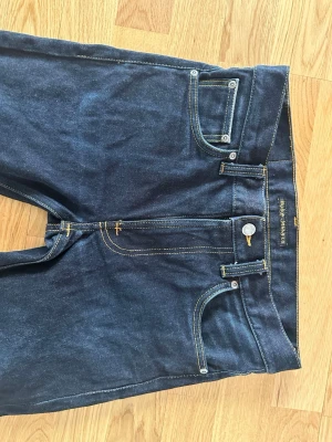 Mörkblå nudie jeans  - Snygga mörkblå nudie jeans med klassisk femficksdesign och raka ben. Jeansen har guldfärgade sömmar och är tillverkade i slitstarkt bomullsjeansmaterial. Perfekta för en stilren och tidlös look.