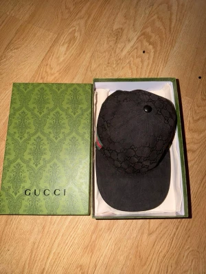 Svart Gucci keps med monogram - Snygg svart keps från Gucci med klassiskt GG-monogram över hela kepsen. Justerbar rem bak och diskret grön-röd rand på sidan. Tillverkad i bomull och polyester, perfekt för dig som vill ha en exklusiv accessoar med streetwear-vibe.