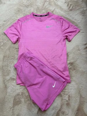 Nike Shortsset - Snyggt rosa träningsset från Nike med t-shirt och shorts. Båda plaggen har Nike-logga i vitt och är tillverkade i ett lätt och snabbtorkande material, perfekt för löpning eller gym. T-shirten har rund hals och kort ärm, shortsen har resår i midjan. T-Shirt storlek S Shorts storlek: M