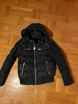 Svart dunjacka från Moose Knuckles - Snygg svart dunjacka från Moose Knuckles med huva och silvriga dragkedjor. Jackan har quiltad design, flera fickor och en modern passform. Perfekt för kalla dagar och ger en cool streetwear-look. Det är original jacka. Pris går absolut att diskutera! Nypris 9000kr