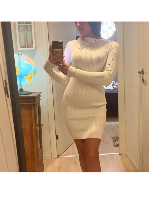 Vit ribbad stickad kortklänning - Snygg vit stickad kortklänning med ribbad struktur och lång ärm. Klänningen har en tight passform och rund halsringning med dekorativa knappar på axeln. Perfekt för dig som gillar en stilren och enkel look.