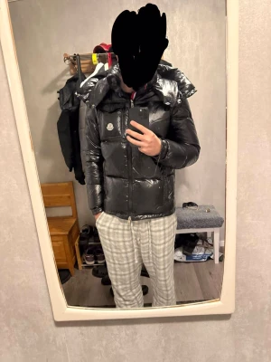 Svart dunjacka från Moncler - Snygg svart dunjacka från Moncler med glansig finish och klassisk logga på bröstet. Jackan har quiltad design, dragkedja framtill och praktisk bröstficka med lock. Perfekt för kalla dagar och ger en riktigt trendig look. Pris kan diskuteras.