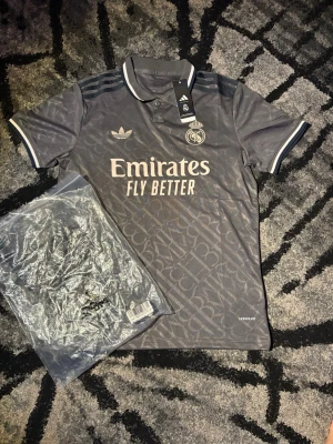 Real Madrid x Adidas grå matchtröja - Snygg grå Real Madrid matchtröja från Adidas med klassisk krage, svarta detaljer och tryckta loggor. Tröjan har mönstrad yta, tre ränder på axlarna och 'Emirates Fly Better' tryckt på bröstet. Perfekt för fotbollsträning eller supporterliv.