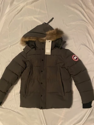 Mörkgrå dunjacka från Canada Goose - Snygg mörkgrå dunjacka från Canada Goose med avtagbar huva och fuskpälsdetalj. Jackan har klassisk pufferstil, dragkedja och knappar framtill samt den ikoniska loggan på ärmen. Perfekt för kalla vinterdagar.