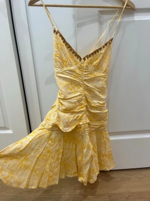 Gul mönstrad kortklänning med volang - Superfin gul kortklänning med vitt mönster och smala axelband som korsas i ryggen. Klänningen har volangkant nertill, rynkade detaljer framtill och en v-ringad hals med små dekorativa bollar. Perfekt för varma dagar och midsommar! Använd ungefär 3-4 gånger 