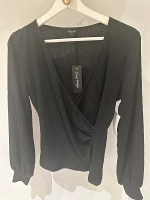 Svart omlottopp från Happy Holly storlek 36/38 - Snygg svart omlottopp från Happy Holly med lätt glittrig finish. Toppen har v-ringning, långa ärmar. Perfekt för dig som vill ha en trendig och elegant look.